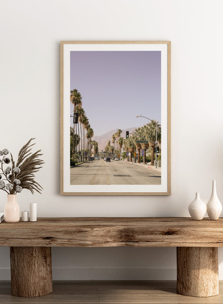 A framed art print featuring a scenic photograph of a palm-lined street in Palm Springs with mountains in the background. Palm-Lined Avenue by Shot by Clint travel photography print featuring a California road, tall palm trees, desert mountains, and soft sun-faded tones.