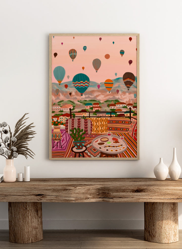 A colorful art print depicting a scenic view of a hillside town with mountains in the background, filled with hot air balloons. In the foreground, a patterned outdoor seating area features a table with food and a potted plant. Shop Balloons Over Rooftop Dinner by Nidhi Kachhadiya, a colorful travel wall art print featuring hot air balloons, rooftop dining, patterned textiles, and teal and coral tones.