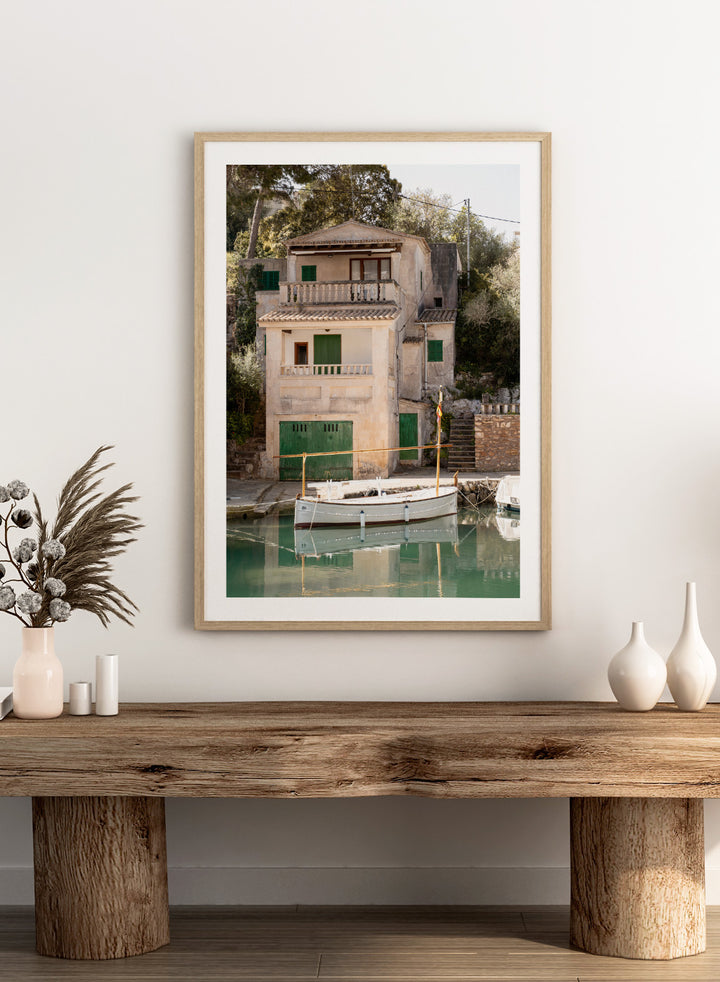 Two-story stone building with green shutters by a body of water with boats. Mediterranean Boathouse by Henrike Schenk is a coastal photography print featuring a waterfront house, fishing boat, turquoise water, and Mediterranean charm.