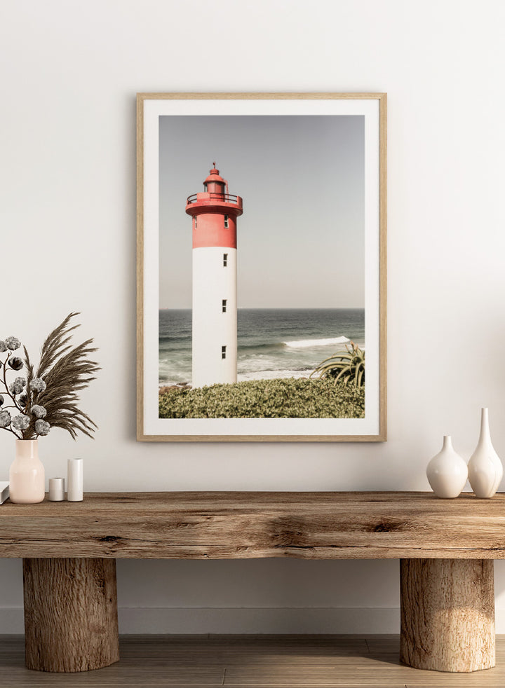 A framed photographic art print of a red and white lighthouse standing on a grassy shore with the ocean and sky in the background. Beacon by the Sea by Shot by Clint coastal photography print featuring a red-and-white lighthouse, ocean waves, pale sky, and coastal greenery.
