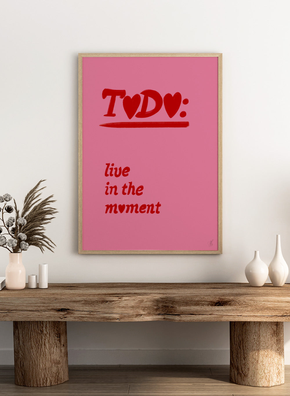 Pink poster with red text 'TODO: live in the moment' on a pink background. Typography wall art reading “live in the moment” with red hearts on pink background by Anne-Marie Volfova. Officially licensed motivational art print.