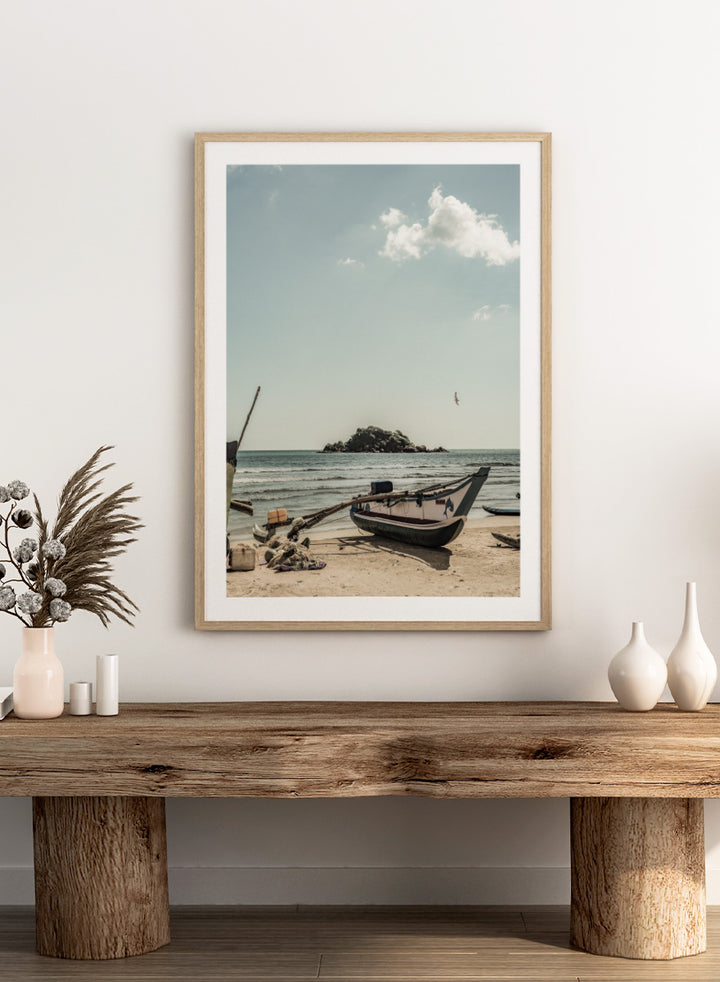 A framed photographic print of a coastal beach scene with a small boat on the sandy shore, the sea and distant rocky island, and a pale blue sky with clouds. Seaside Serenity by Shot by Clint coastal photography print featuring a fishing boat, sandy shore, ocean waves, distant island, and weathered seaside details.
