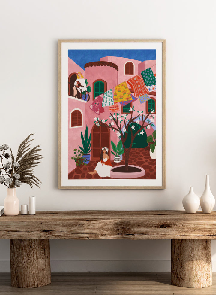 A framed art print featuring a colorful Mediterranean-style illustration of a pink courtyard with a woman sitting under a flowering tree, laundry hanging in the background, and a blue sky above. Pink Village Courtyard by Ceyda Alasar art print featuring pink architecture, patterned rugs, flowering trees, potted plants, and relaxed courtyard life.