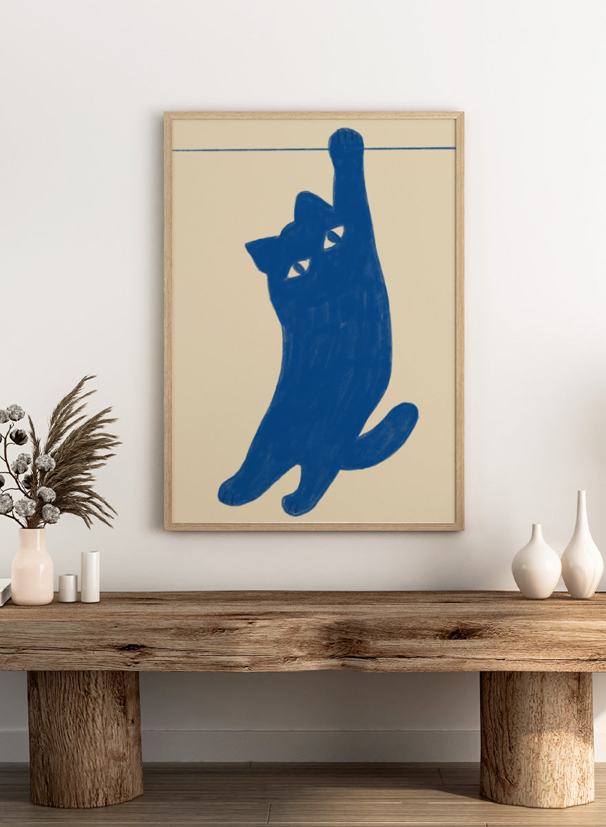 A stylized blue cat silhouette hanging from a thin horizontal line on a warm beige background. Hanging Blue Cat by The Art Concept is a playful cat art print featuring a blue cat dangling from one paw against a warm cream background.