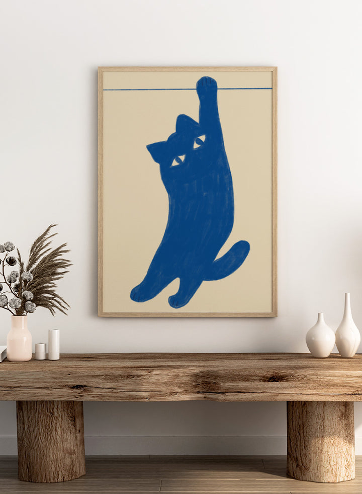 A stylized blue cat silhouette hanging from a thin horizontal line on a warm beige background. Hanging Blue Cat by The Art Concept is a playful cat art print featuring a blue cat dangling from one paw against a warm cream background.