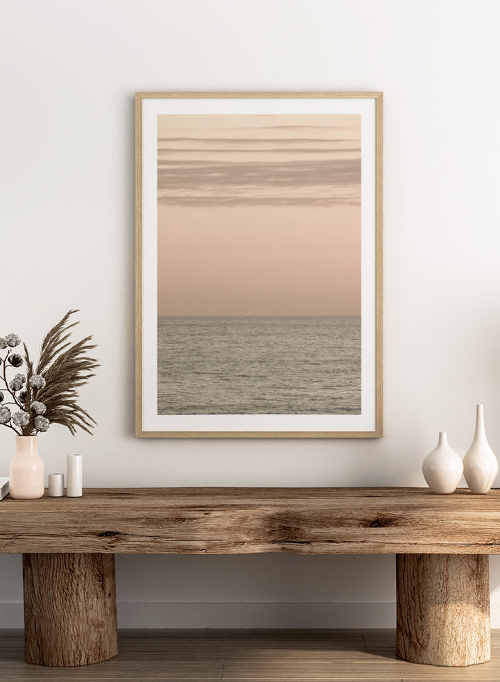 A minimalist framed art print depicting a calm seascape at dusk or dawn, with a soft peach and beige sky meeting a muted gray-blue ocean horizon. Serene Horizons by Shot by Clint features a soft peach sky and calm ocean horizon in a minimal coastal photograph with airy neutral tones.