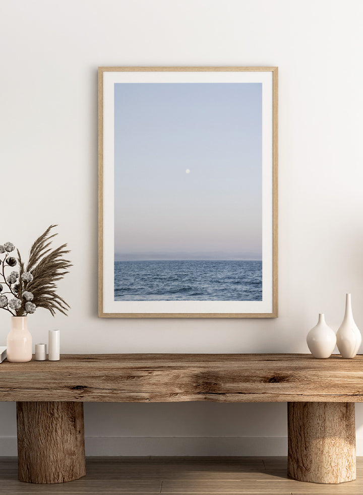 A minimalist framed art print depicting a calm blue ocean under a pale sky with a small visible moon. Moonlit Horizon by Shot by Clint coastal photography print featuring a pale moon, distant shoreline, open blue water, and soft lavender evening tones.