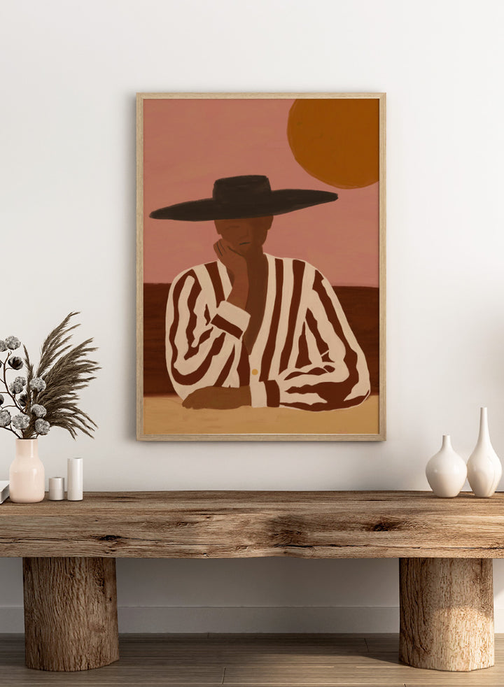A stylized art print depicting a seated figure in a wide-brimmed black hat and a brown-and-white vertically striped shirt, set against a dusty pink sky with a large orange sun. Desert Dreamer by The Art Concept art print featuring a relaxed figure in a wide black hat, striped shirt, dusty pink sky, and warm desert-inspired tones.