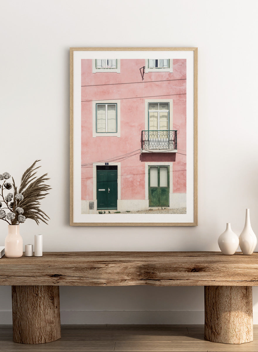 A framed art print featuring a photograph of a pink building facade with green doors, white-framed windows, and a small black metal balcony. Pink Façade Harmony by Shot by Clint architectural photography print featuring a pink exterior, green doors, white windows, and an iron balcony.