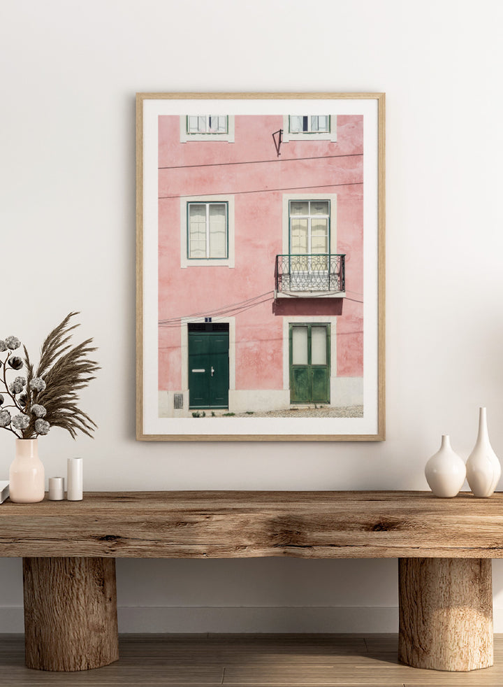 A framed art print featuring a photograph of a pink building facade with green doors, white-framed windows, and a small black metal balcony. Pink Façade Harmony by Shot by Clint architectural photography print featuring a pink exterior, green doors, white windows, and an iron balcony.