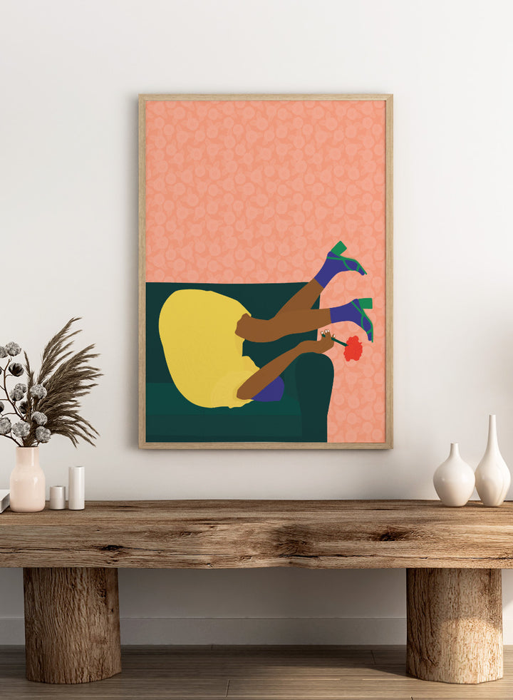 Illustration of a person sitting on a couch with a flower, against a patterned pink background. New Year’s Eve by Jota De Jai is a playful modern fine art print of a woman upside down on a couch in heels and a yellow dress. Available framed or unframed.