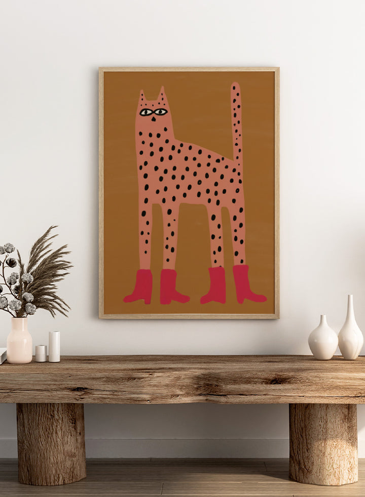A stylized art print of a tall spotted cat in a pinkish-orange tone with black spots and bright pink boots, set against a warm brown background. Spotted Cat in Boots by The Art Concept playful cat art print featuring a pink spotted cat with long legs, red boots, and a warm ochre background.