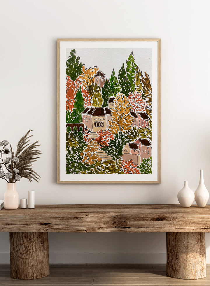 A framed art print featuring a hand-drawn, stylized landscape of a hillside village with beige buildings, brown roofs, and dense trees in green, yellow, orange, and red tones. Shop Autumn Hillside Village by Rushati Chowdhury, a colorful autumn landscape art print with hillside homes, winding steps, dense trees, and warm fall tones.