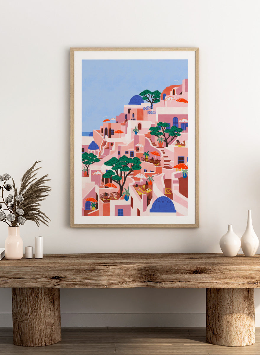 A stylized art print depicting a hillside Mediterranean village with pink and peach buildings, blue domes, green trees, and a light blue sky. Santorini Summer Dream by Ceyda Alasar art print featuring blue domes, terraced Mediterranean architecture, orange umbrellas, and sunny Greek island charm.