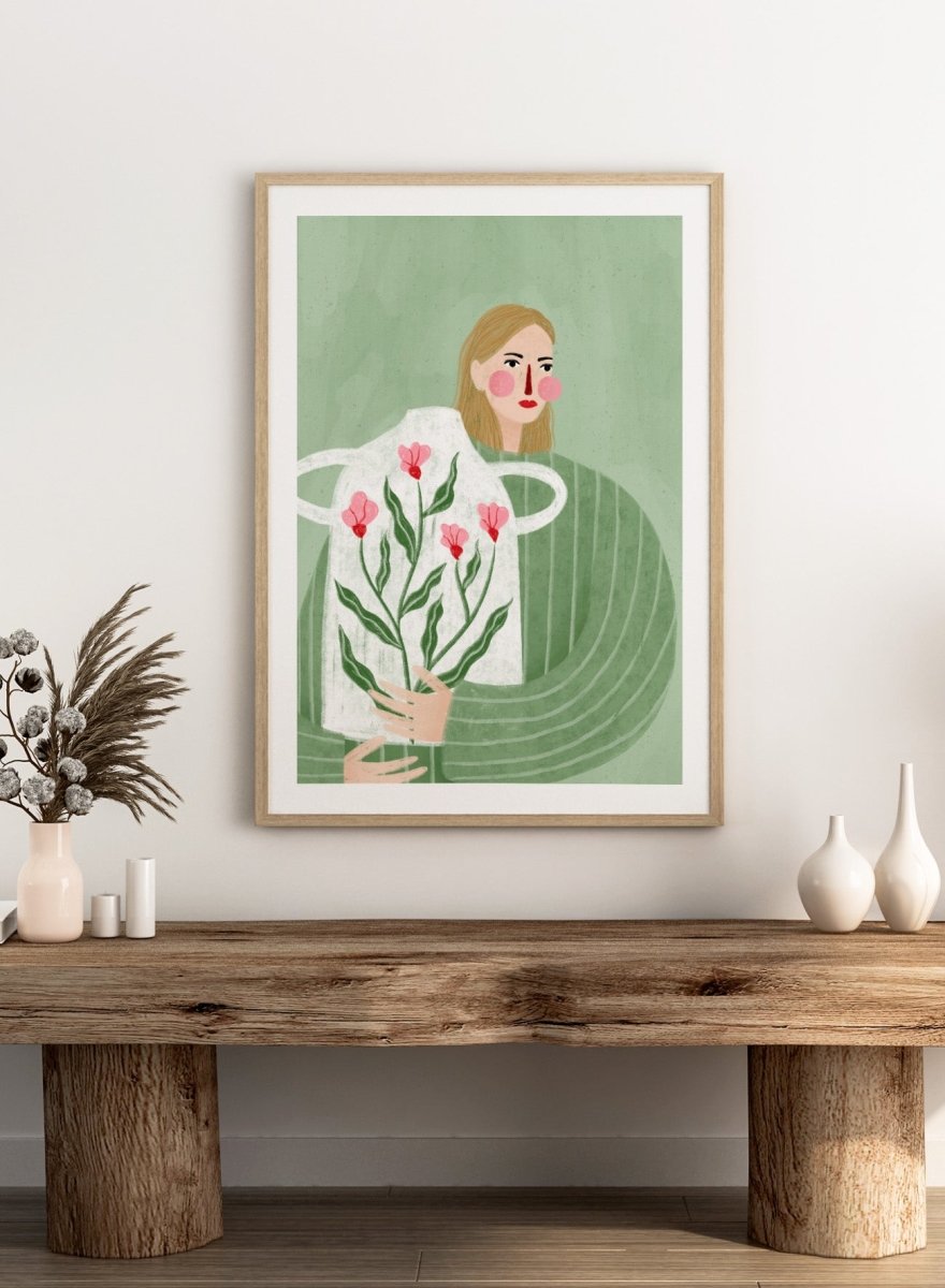A framed art print featuring a stylized illustration of a woman with blonde hair holding a white apron decorated with pink flowers and green leaves, set against a sage green background. Woman Holding Flowers by Bea Müller premium framed fine art print featuring a woman carrying a white vase filled with pink flowers and green leaves.
