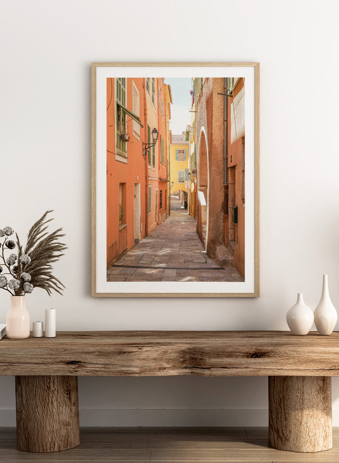 Framed photograph of a narrow street on a wall next to a window.  narrow street with colorful buildings in a European city. Vibrant European Alley by Henrike Schenk is a travel photography print featuring a Mediterranean street, cobblestone alley, and pastel European architecture.