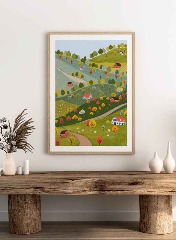A framed art print featuring a whimsical, stylized illustration of a rolling green and yellow countryside landscape with red houses, colorful autumn trees, winding roads, and a pale blue sky. Vibrant Autumn Village Hills by Ceyda Alasar art print with rolling countryside, colorful fall trees, farmhouses, barns, sheep, and winding roads.