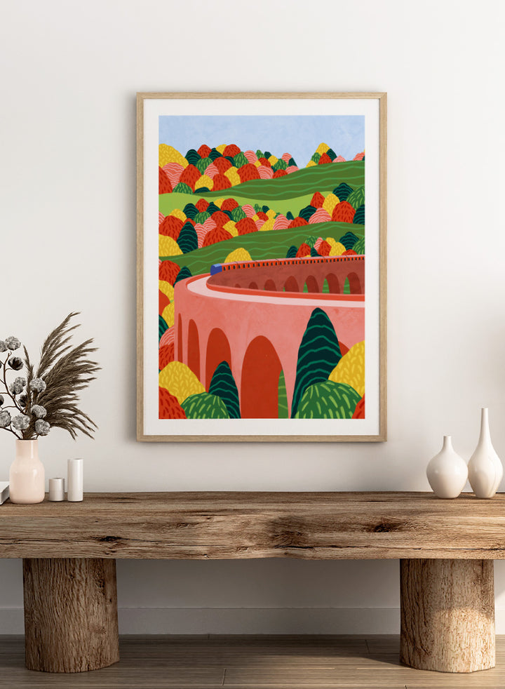 A framed art print featuring a stylized landscape illustration with rolling green hills, colorful patterned trees, and a pink arch bridge under a light blue sky. Winding Autumn Viaduct by Ceyda Alasar travel art print featuring a colorful train, arched bridge, rolling hills, and bold autumn foliage.