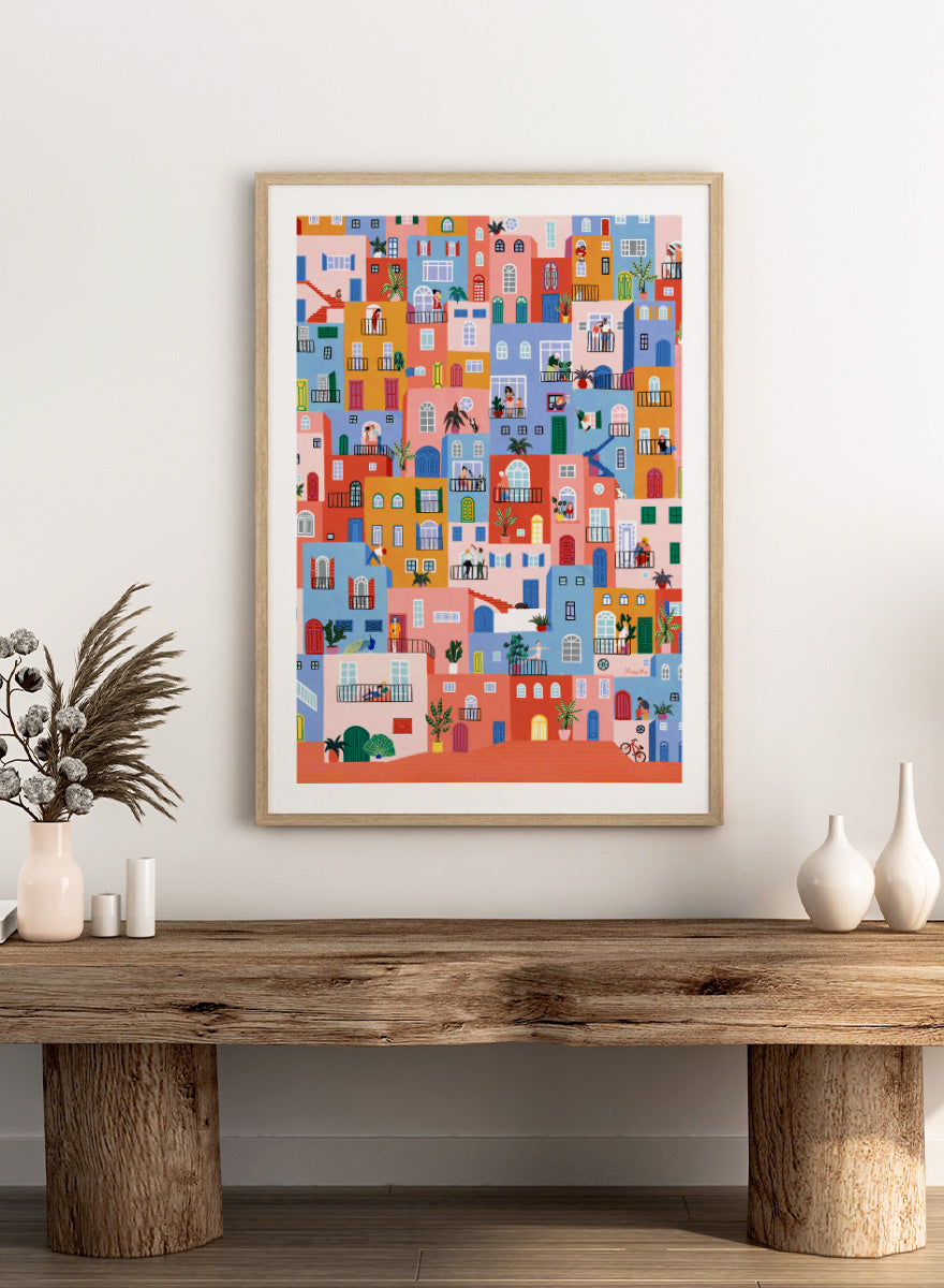 A framed art print featuring a colorful, whimsical illustration of Mediterranean-style buildings with people, animals, and plants. Colorful Town Life by Ceyda Alasar city art print featuring vibrant buildings, balconies, neighbors, plants, and lively urban details in bold color.