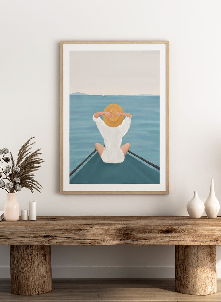 Person sitting on a dock over water wearing a white dress and straw hat. Ocean Escape by Flow Line features a woman sitting on a boat overlooking the sea, creating a coastal art print inspired by travel, summer, and ocean living.