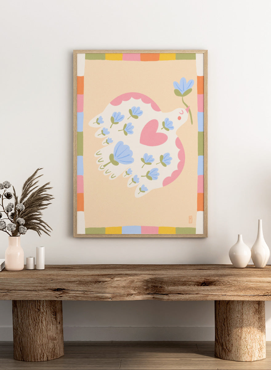 A framed art print featuring a whimsical illustration of a white dove with blue flowers and a pink heart, framed by a pastel multicolor border on a light peach background. Shop Flowering Peace Dove by Dirkie Nell, a colorful bird and floral art print featuring a white dove, blue flowers, pink wings, and a playful peace motif.