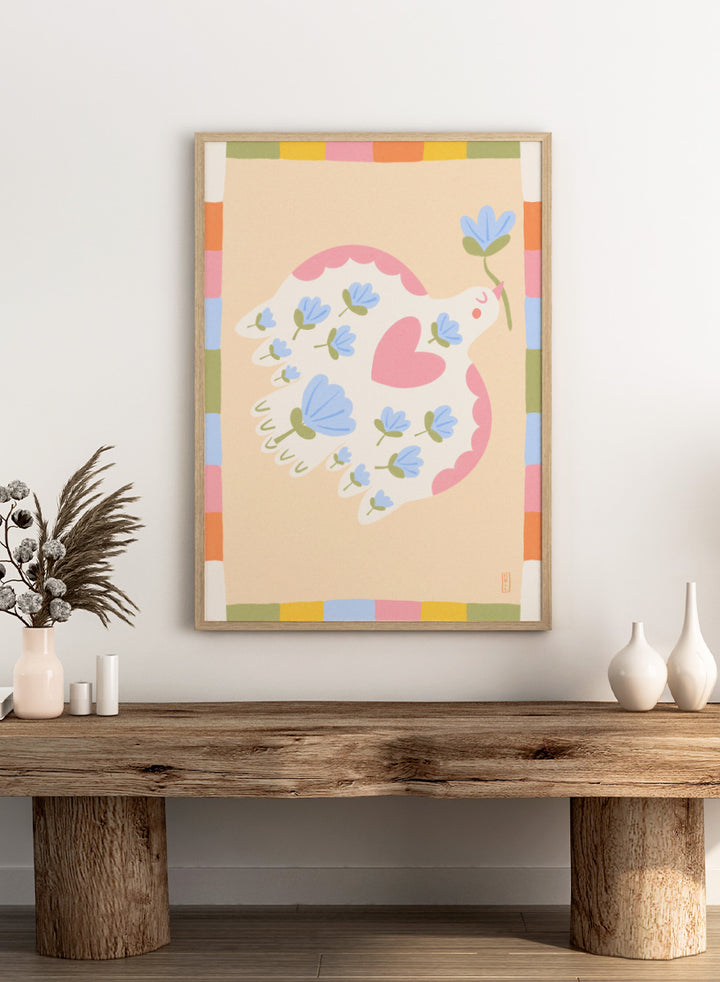 A framed art print featuring a whimsical illustration of a white dove with blue flowers and a pink heart, framed by a pastel multicolor border on a light peach background. Shop Flowering Peace Dove by Dirkie Nell, a colorful bird and floral art print featuring a white dove, blue flowers, pink wings, and a playful peace motif.