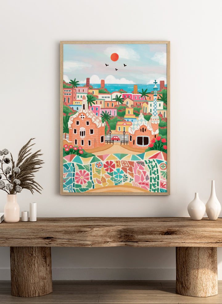 A colorful stylized illustration of a coastal town with pastel buildings, palm trees, a mosaic wall in the foreground, blue sea, and a red sun in the sky. Shop Colorful Coastal Dream by Nidhi Kachhadiya, a vibrant Barcelona wall art print inspired by Park Güell, Gaudí architecture, mosaics, and bright coastal color.