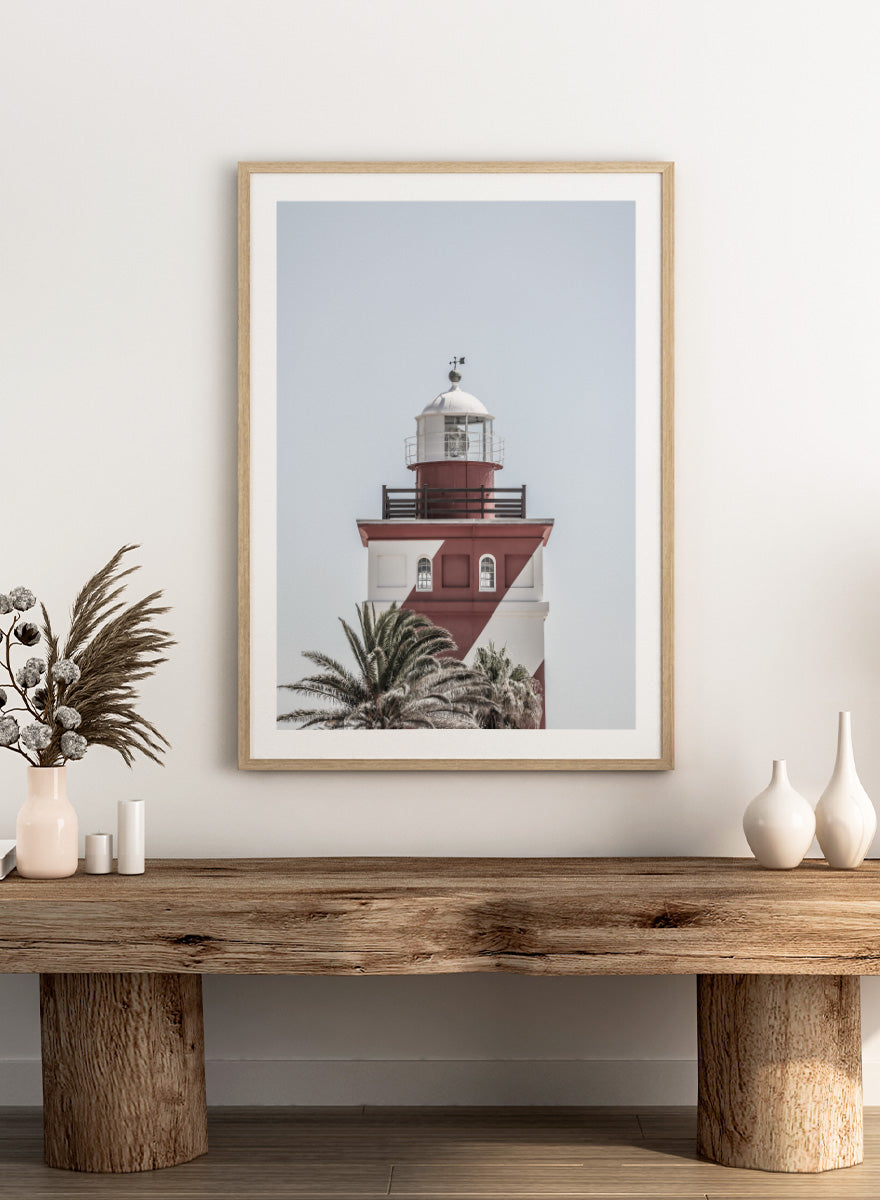 A framed art print featuring a red and white lighthouse with palm trees at the bottom, set against a pale sky background. Beacon of Solitude by Shot by Clint lighthouse photography print featuring red-and-white architecture, palm trees, arched windows, and a pale coastal sky.