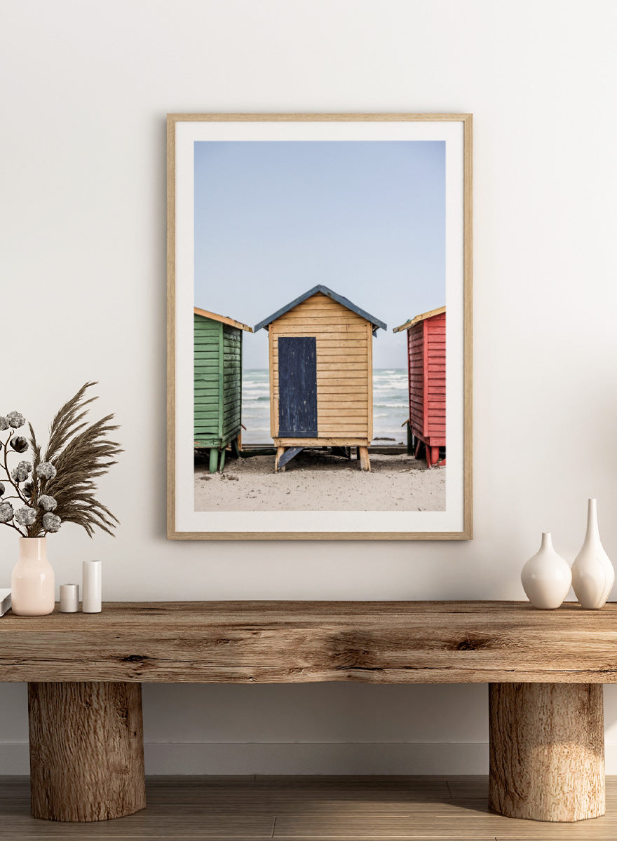 A framed photographic print of three colorful wooden beach huts on a sandy beach, with the central hut in natural wood and a dark blue door, flanked by green and red huts under a pale blue sky. Colorful Beach Huts by Shot by Clint coastal photography print featuring green, natural wood, and red huts with weathered texture and ocean views.