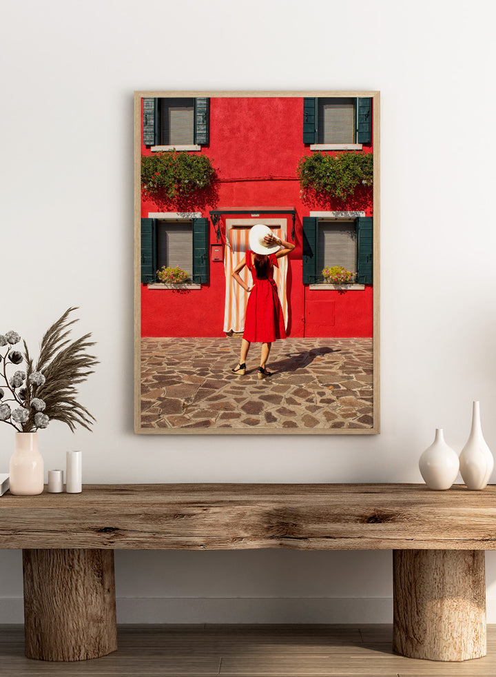 Woman in a red dress and white hat standing in front of a red wall with green shutters. Red Art Print by Anette Ohlendorf featuring a woman in a red dress standing against a red building. Officially licensed travel photography with strong color and symmetry.