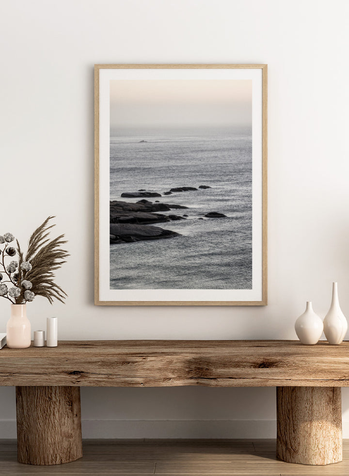 A framed black and white coastal landscape photograph featuring a calm sea, distant rocks, and a pale sky. Silver Sands by Shot by Clint coastal photography print featuring silver-blue water, dark shoreline rocks, and a soft misty horizon.