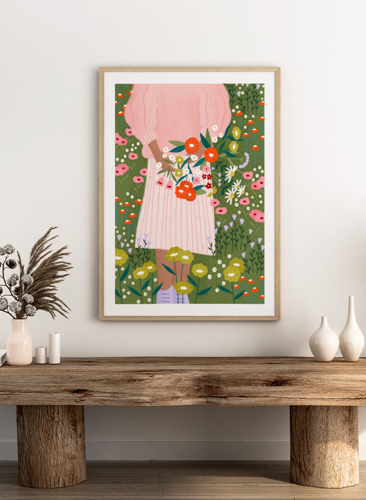 A framed art print featuring a stylized illustration of a person in a pink striped dress holding a bouquet of orange, red, and white flowers in a green field of wildflowers. Secret Garden Bouquet by Bea Müller floral art print featuring a woman gathering wildflowers in a colorful garden filled with poppies, daisies, and greenery.