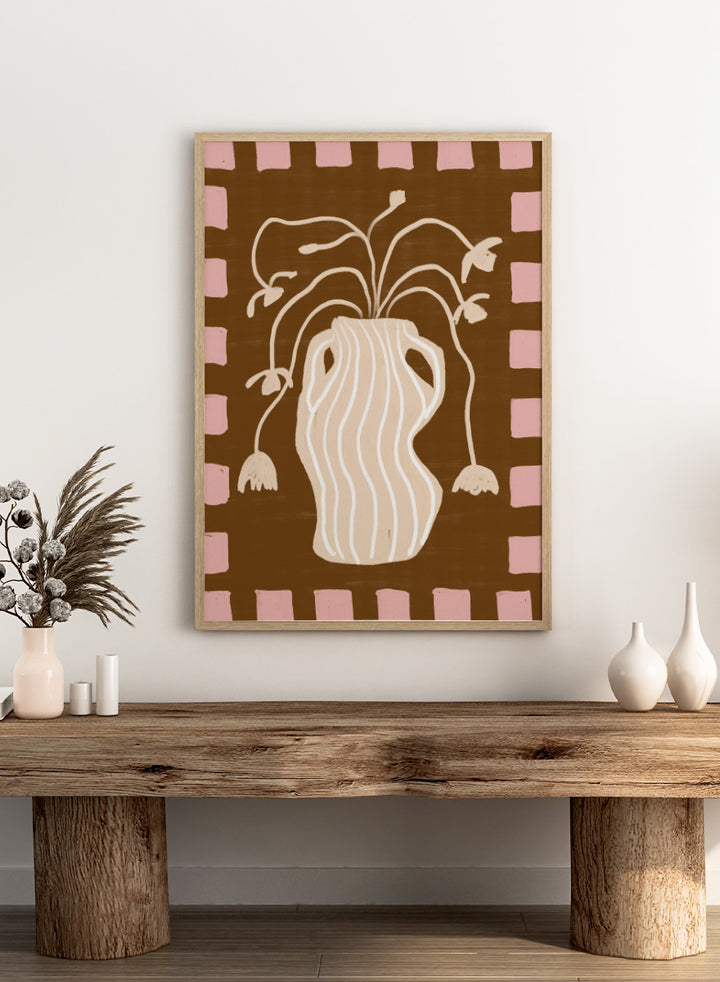 A stylized art print featuring a cream-colored wavy vase with stems and hanging flower buds on a brown background, framed by a pink and brown checkered border. Soft Floral Arrangement by The Art Concept still life art print featuring a striped vase, drooping flowers, warm brown tones, and a soft pink border.