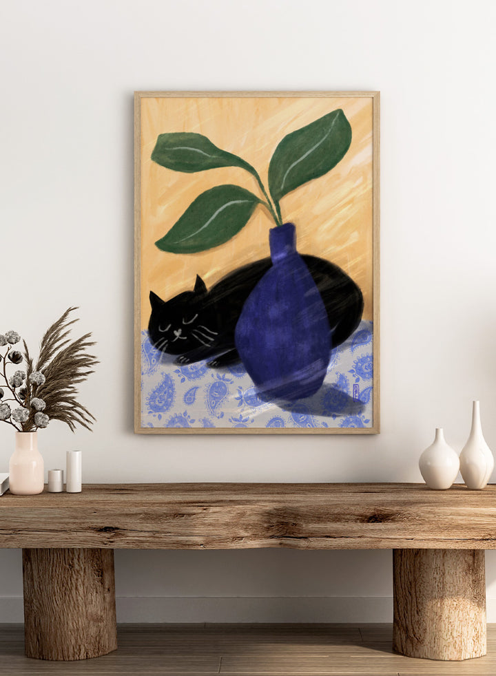 A stylized still life painting featuring a black cat lying on a blue-and-white patterned surface beside a dark blue vase with green leaves, against a warm beige background. Shop the Sleeping Black Cat art print by Dirkie Nell, featuring a curled black cat, bold cobalt blue vase, green leaves and playful patterned details.