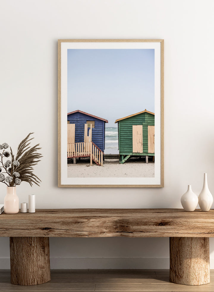 A framed photographic print of two wooden beach huts on a sandy beach under a pale blue sky, with one hut in dark blue and the other in green, both with light wood shutters and doors. Coastal Huts Harmony by Shot by Clint coastal photography print featuring blue and green beach huts, sandy ground, weathered wood, and ocean views.