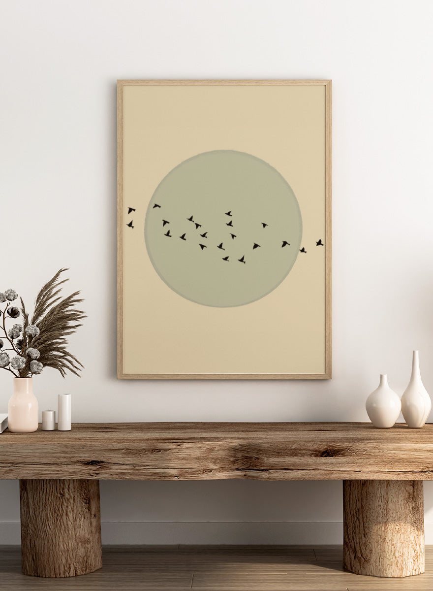 Minimalist art print featuring a large muted sage green circle centered on a warm beige background, with small black bird silhouettes flying around it. Flock Against The Moon by The Art Concept is a minimalist bird art print featuring black birds in flight across a pale green moon and warm cream background.
