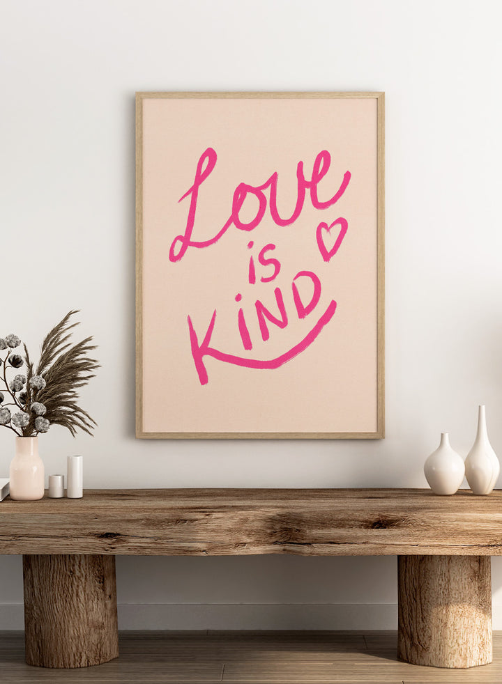 Pink text 'Love is Kind' with a heart on a light pink background. Love Is Kind by Dirkie Nell is a pink typography art print with handwritten lettering and heart detail. A joyful modern love wall poster.