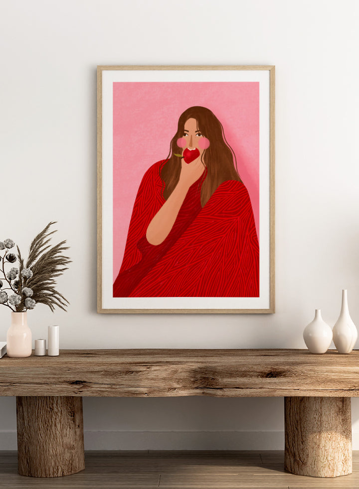 A framed art print featuring a stylized illustration of a woman with long brown hair, wearing a red patterned wrap, holding a red apple to her lips, set against a pink background. Cozy Woman Apple by Bea Müller portrait art print featuring a woman eating a red apple in bold red and pink tones for colorful modern wall decor.