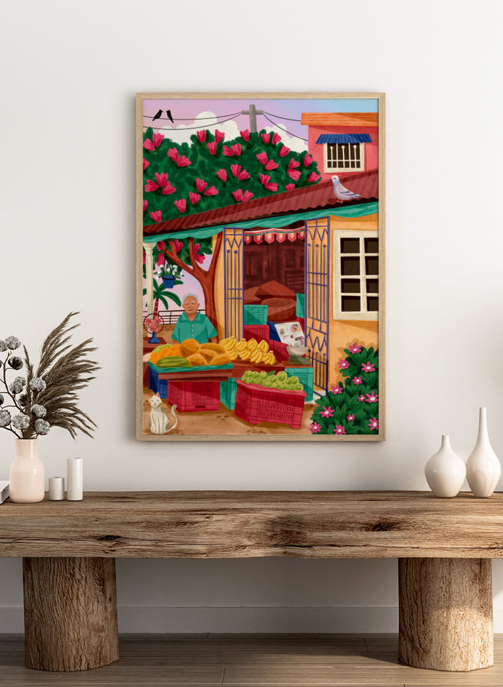 A colorful illustrated street market scene with a vendor standing behind tables of fruit and vegetables, a flowering tree, a house with pink flowers, crates, a pigeon on the roof, birds in the sky, and a white cat in the lower left. Colorful fruit stand art print by Nidhi Kachhadiya with fresh produce, floral details, and market charm. A cheerful print for kitchen or dining spaces.