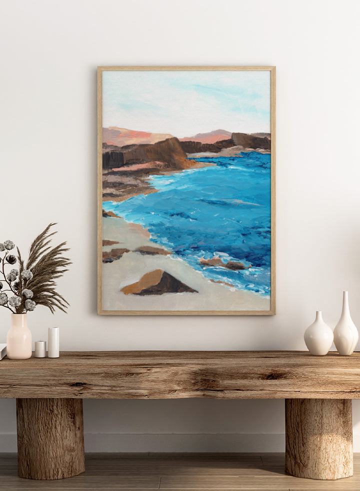 A framed landscape painting depicting a coastal scene with bright blue ocean water, sandy shore, dark rocky cliffs, and distant hills under a pale sky. Shop Rocky Coastal Waters by Nikita Jariwala, a coastal wall art print poster with blue ocean water, rocky cliffs, and a peaceful seaside view.