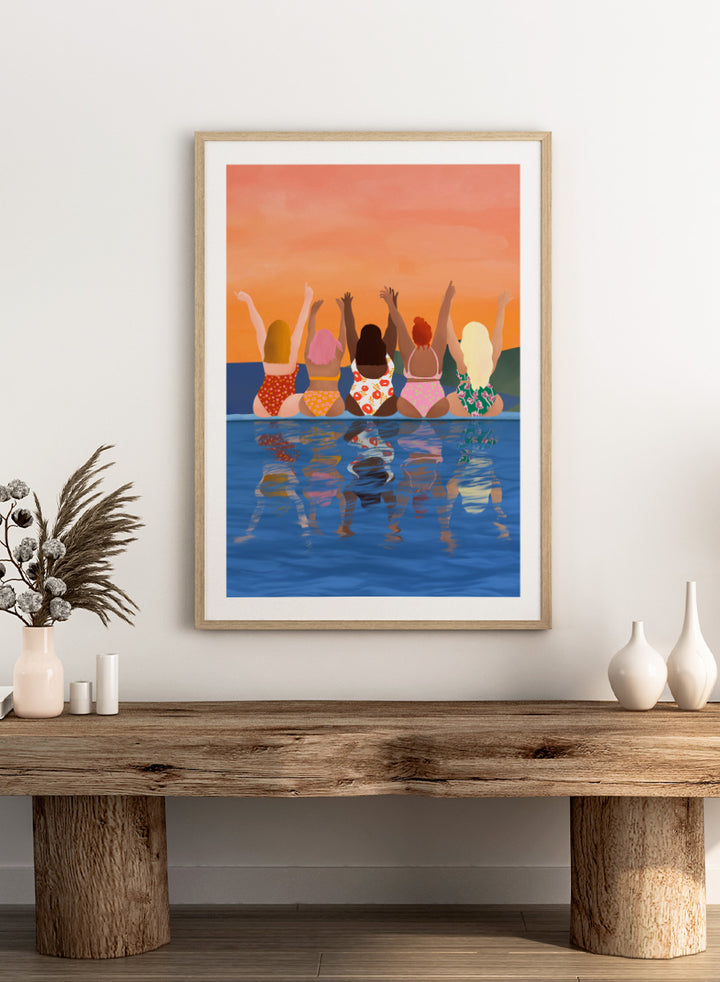 A framed art print featuring a stylized illustration of five women sitting in a pool at sunset, seen from behind as they raise their arms. The background is a warm orange sky, and the water below reflects their silhouettes. Summer Sunset Friends by Ceyda Alasar friendship art print featuring five women by the water in patterned swimsuits beneath a glowing orange sky.