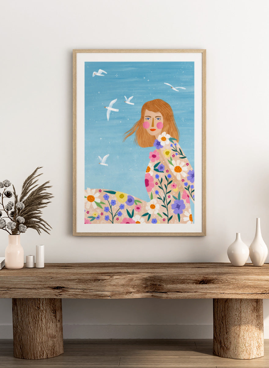 A framed art print featuring a stylized illustration of a young woman with long light brown hair, surrounded by colorful flowers and white birds against a light blue sky. ree Spirit Floral Dreams by Bea Müller floral woman art print featuring a flowing flower-covered summer dress, white birds, and a bright blue sky.