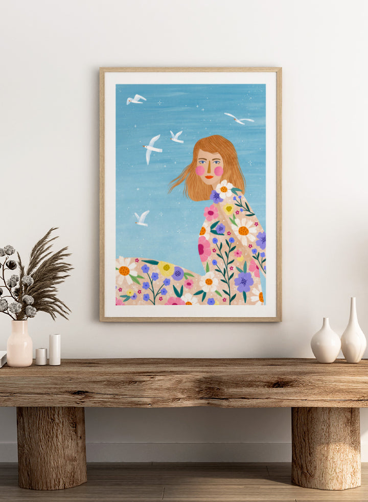 A framed art print featuring a stylized illustration of a young woman with long light brown hair, surrounded by colorful flowers and white birds against a light blue sky. ree Spirit Floral Dreams by Bea Müller floral woman art print featuring a flowing flower-covered summer dress, white birds, and a bright blue sky.