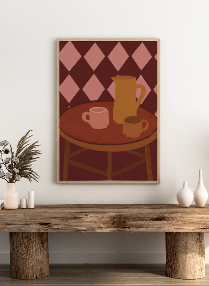 A stylized illustration of a round wooden table with a pink mug, a yellow pitcher, and a small cup against a dark red and pink diamond-patterned background. Retro Coffee Time by The Art Concept warm art print featuring mugs, a pitcher, and a café table in soft retro tones and a diamond pattern background.