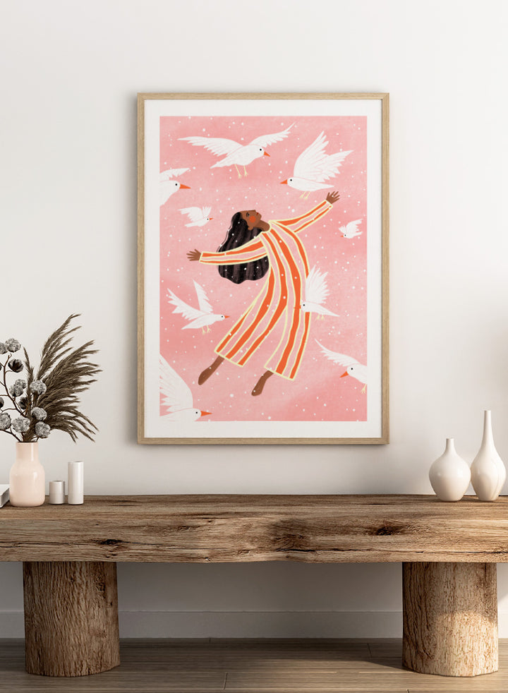 A framed art print featuring an illustration of a woman in orange and white striped pajamas floating among white doves on a soft pink background. Joyful Liberation by Bea Müller woman art print featuring a free-spirited figure surrounded by white birds in bold pink and orange tones.