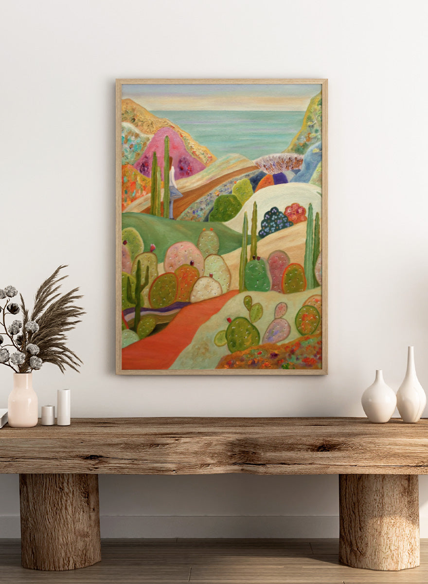A colorful landscape painting featuring stylized cacti and succulents in a desert valley, with rolling hills, a pink mountain, and a blue sea or sky in the background. Shop Colorful Desert Yoga View by Angeles M. Pomata, a colorful desert landscape art print featuring cacti, rolling hills, a yoga figure, and coastal views.