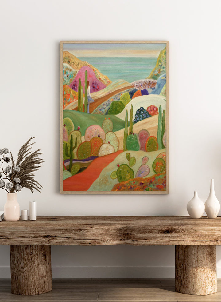 A colorful landscape painting featuring stylized cacti and succulents in a desert valley, with rolling hills, a pink mountain, and a blue sea or sky in the background. Shop Colorful Desert Yoga View by Angeles M. Pomata, a colorful desert landscape art print featuring cacti, rolling hills, a yoga figure, and coastal views.