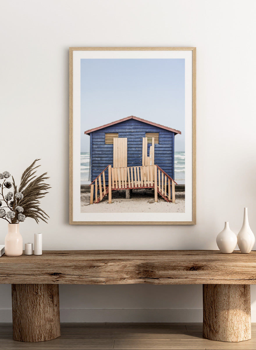 A framed photographic print of a blue wooden beach lifeguard hut on a sandy shore with the ocean and pale sky in the background. Beachside Retreat by Shot by Clint features a weathered blue beach hut and soft ocean backdrop in a calm coastal photography print.