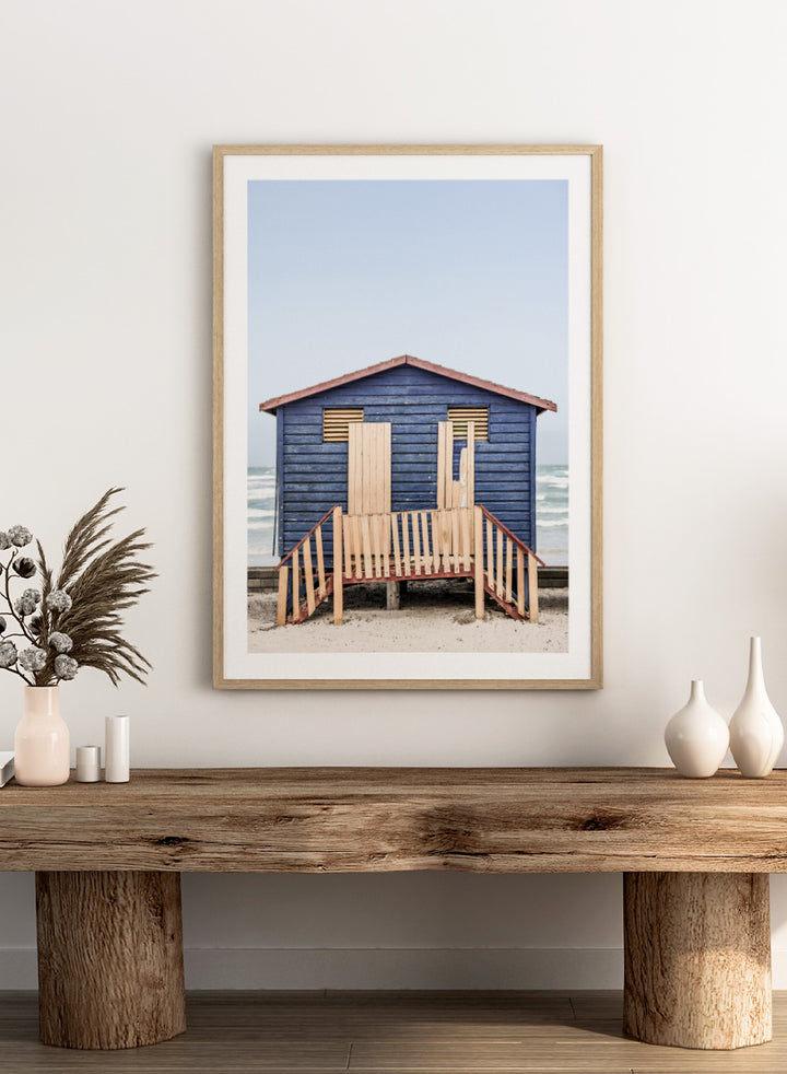 A framed photographic print of a blue wooden beach lifeguard hut on a sandy shore with the ocean and pale sky in the background. Beachside Retreat by Shot by Clint features a weathered blue beach hut and soft ocean backdrop in a calm coastal photography print.