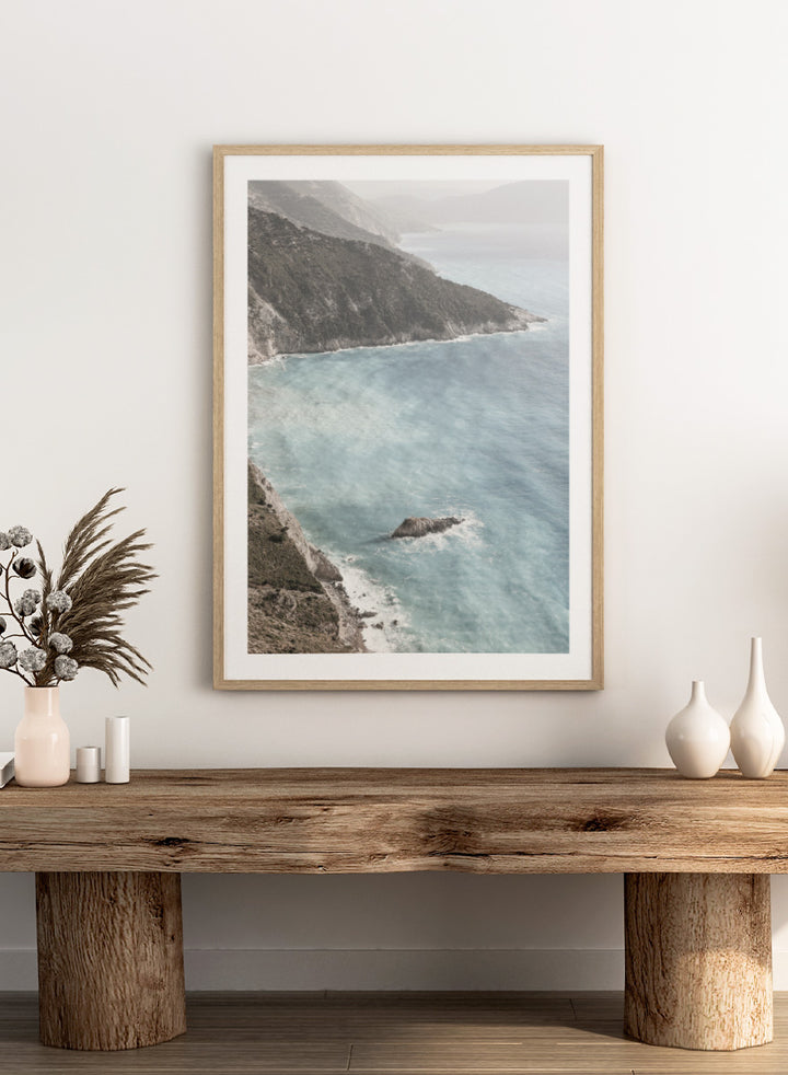 A framed photographic art print depicting a coastal landscape with a steep rocky cliff overlooking a calm, pale blue sea with a small rock formation in the foreground. Coastal Serenity by Shot by Clint coastal photography print featuring pale blue water, rugged cliffs, rocky shores, and a soft hazy horizon.